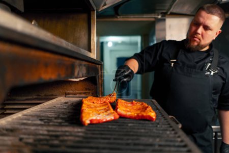 Foto de En una cocina profesional un chef en una chaqueta negra pone costillas marinadas cerca de un horno a la parrilla caliente - Imagen libre de derechos