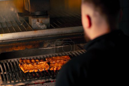 Foto de Primer plano de un chef de cocina profesional en una chaqueta negra cerca de un horno a la parrilla caliente gira sobre las costillas - Imagen libre de derechos