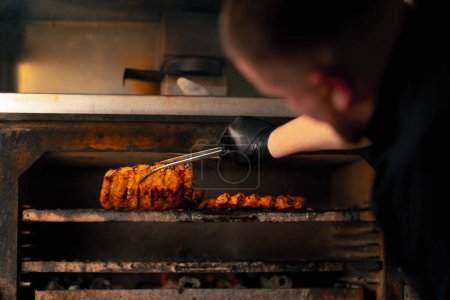 Photo for Close-up of a professional kitchen chef in a black jacket near a hot grill oven turns over ribs - Royalty Free Image