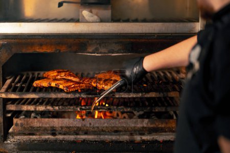 Photo for Close-up of a professional kitchen chef in a black jacket near a hot grill oven turns over ribs - Royalty Free Image