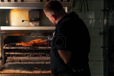Photo for A professional kitchen chef in a black jacket near a hot grill oven turns over ribs - Royalty Free Image