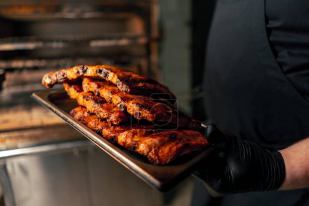 Photo for Close-up of a professional kitchen a chef in a black jacket near a hot grill oven takes ribs out of the grill - Royalty Free Image