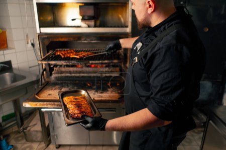 Foto de Una cocina profesional un chef en una chaqueta negra cerca de un horno a la parrilla caliente saca las costillas de la parrilla - Imagen libre de derechos