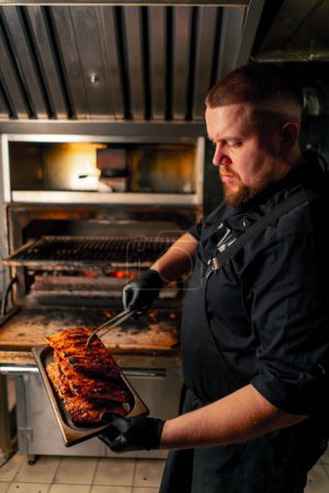 Téléchargez les photos : Une cuisine professionnelle un chef vêtu d'une veste noire près d'un four à barbecue chaud sort les côtes levées du gril - en image libre de droit