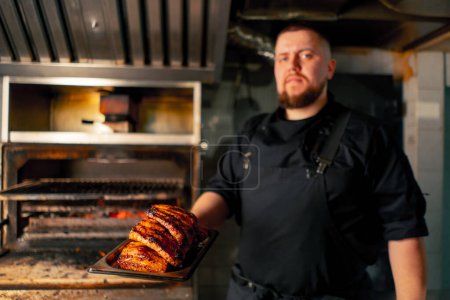 Photo for A professional kitchen chef near a hot grill oven with ribs looking at the camera - Royalty Free Image