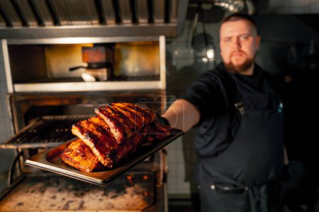 Foto de Un chef de cocina profesional cerca de un horno a la parrilla caliente con costillas mirando a la cámara - Imagen libre de derechos