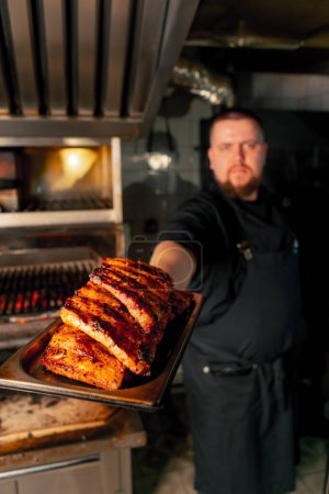 Photo for A professional kitchen chef near a hot grill oven with ribs looking at the camera - Royalty Free Image