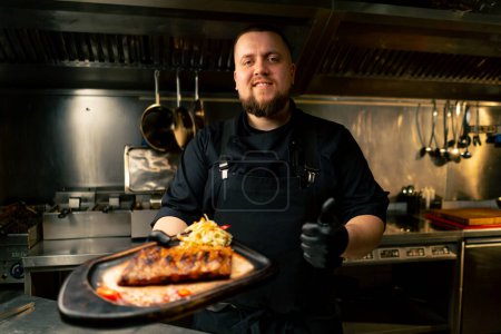 Téléchargez les photos : Dans la cuisine professionnelle, le chef se tient dans ses mains avec un plat préparé et sourit à la caméra - en image libre de droit
