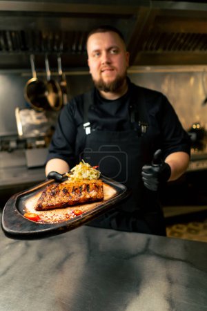 Foto de En la cocina profesional el chef está en sus manos con un plato preparado y sonríe a la cámara - Imagen libre de derechos