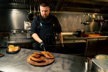 Foto de En la cocina profesional el chef se para cerca de la mesa y plancha las costillas sobre el tablero con pinzas - Imagen libre de derechos