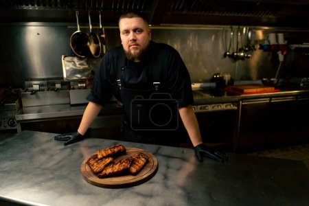 Foto de En la cocina profesional el chef se para cerca de la mesa con las costillas mirando a la cámara - Imagen libre de derechos