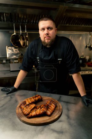 Foto de En la cocina profesional el chef se para cerca de la mesa con las costillas mirando a la cámara - Imagen libre de derechos