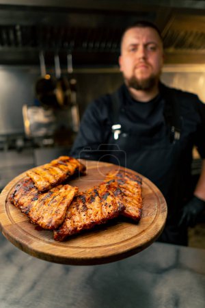 Photo for In professional kitchen the chef holds a board with ribs and demonstrates looking at the camera - Royalty Free Image