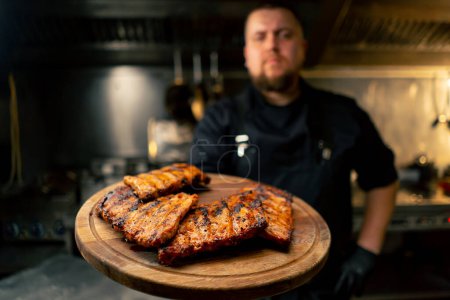 Photo for In professional kitchen the chef holds a board with ribs and demonstrates looking at the camera - Royalty Free Image