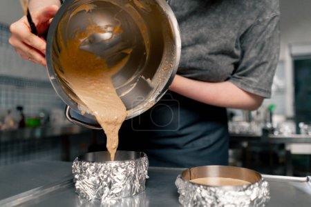 Foto de Cerrar las manos panadero femenino en una cocina profesional vierte masa en capas de moldes de pastel - Imagen libre de derechos