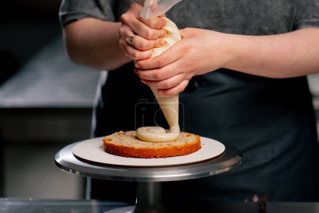 Téléchargez les photos : Fermer boulanger femelle dans une cuisine professionnelle met la crème d'un sac à pâtisserie sur un gâteau éponge - en image libre de droit