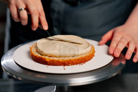 Foto de Primer plano hembra panadero en un profesional cocina distribuye crema en un bizcocho con una espátula - Imagen libre de derechos