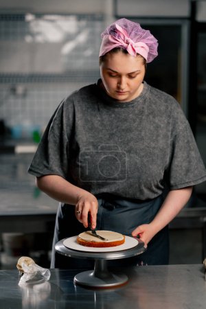 Téléchargez les photos : Femme boulangère dans une cuisine professionnelle distribue de la crème sur un gâteau éponge avec une spatule - en image libre de droit