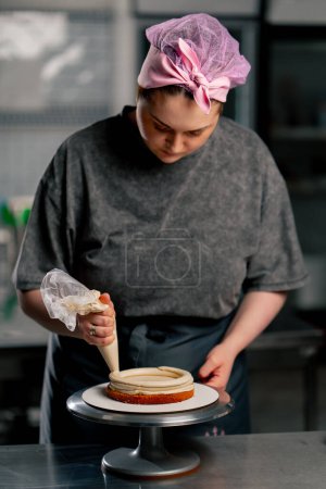 Téléchargez les photos : Femelle boulanger dans une cuisine professionnelle met la crème d'un sac à pâtisserie sur un gâteau éponge - en image libre de droit