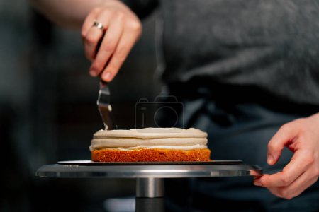 Foto de Primer plano hembra panadero en un profesional cocina distribuye crema en un bizcocho con una espátula - Imagen libre de derechos