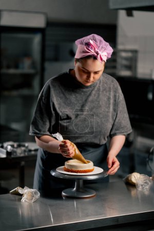 Foto de Panadero femenino en una cocina profesional pone crema de una bolsa de pastelería en un pastel de esponja - Imagen libre de derechos
