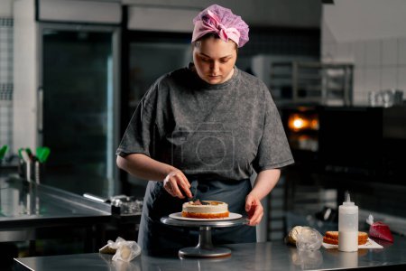 Foto de Panadero femenino en una cocina profesional distribuye crema en un pastel de esponja con una espátula - Imagen libre de derechos