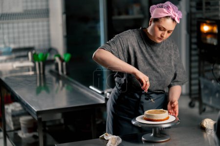 Photo for Female baker in a professional kitchen distributes cream onto a sponge cake with a spatula - Royalty Free Image