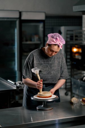 Téléchargez les photos : Fermer boulanger femelle dans une cuisine professionnelle met la crème d'un sac à pâtisserie sur un gâteau éponge - en image libre de droit