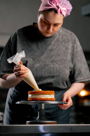 Photo for Female baker in a professional kitchen puts cream from a pastry bag onto a sponge cake - Royalty Free Image