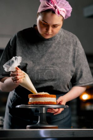 Foto de Panadero femenino en una cocina profesional pone crema de una bolsa de pastelería en un pastel de esponja - Imagen libre de derechos