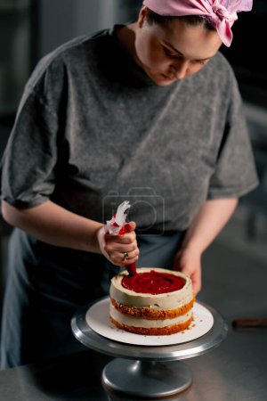Téléchargez les photos : Femelle boulanger dans une cuisine professionnelle met la crème rouge d'un sac à pâtisserie sur un gâteau éponge - en image libre de droit