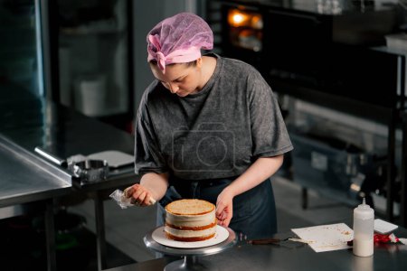 Foto de Panadero femenino en una cocina profesional pone crema roja de una bolsa de pastelería en un pastel de esponja - Imagen libre de derechos