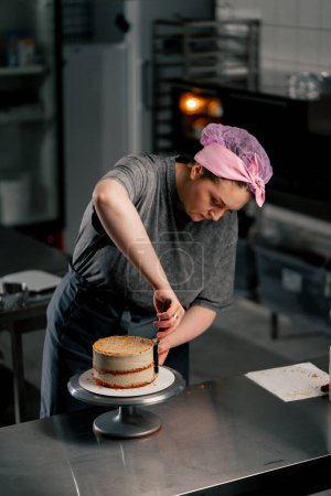 Téléchargez les photos : Femme boulangère dans une cuisine professionnelle distribue de la crème sur un gâteau éponge avec une spatule - en image libre de droit