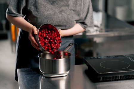 Photo for Close up female baker in a professional kitchen pours frozen cherries into a bowl on a scale - Royalty Free Image