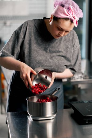 Téléchargez les photos : Femme boulangère dans une cuisine professionnelle verse des cerises congelées dans un bol sur une échelle - en image libre de droit