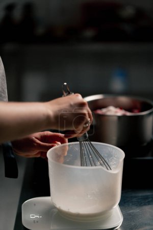 Téléchargez les photos : Gros plan du boulanger dans une cuisine professionnelle fouettant la pâte dans un récipient transparent avec un fouet à la main - en image libre de droit