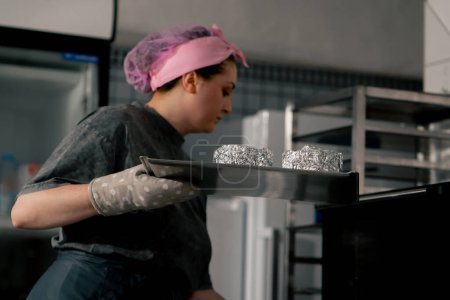 Foto de En una cocina profesional un panadero pone moldes listos en una bandeja para hornear en el horno - Imagen libre de derechos