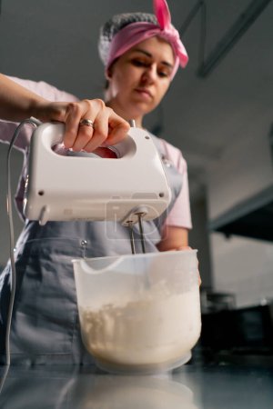 Téléchargez les photos : Plan rapproché d'une boulangère dans une cuisine professionnelle mélangeant crème et colorant avec un mélangeur - en image libre de droit