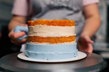 Foto de Cerca de panadero femenino en una cocina profesional pone crema de una bolsa de pastelería en un pastel de esponja - Imagen libre de derechos