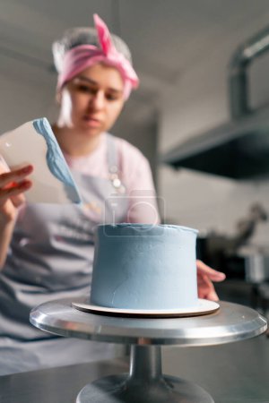 Photo for Close up female baker in a professional kitchen distributes blue cream onto a sponge cake with a spatula - Royalty Free Image