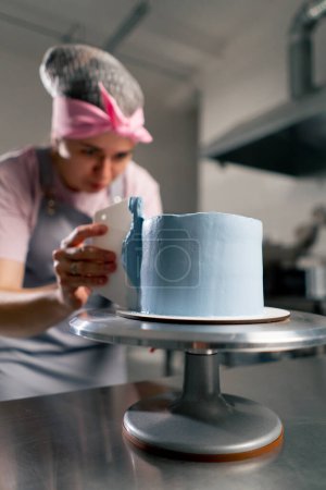 Foto de Primer plano panadero femenino en una cocina profesional distribuye crema azul en un pastel de esponja con una espátula - Imagen libre de derechos