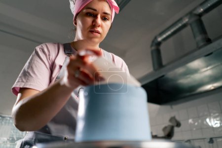 Téléchargez les photos : Gros plan femme boulanger dans une cuisine professionnelle distribue la crème bleue sur un gâteau éponge avec une spatule - en image libre de droit