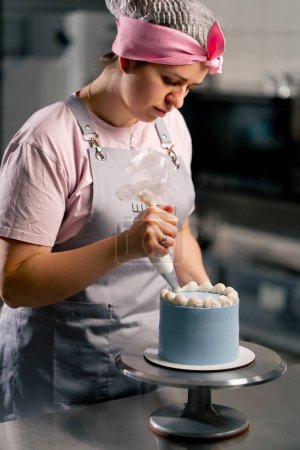 Téléchargez les photos : D'une boulangère dans une cuisine professionnelle décorant un gâteau bleu avec de la crème - en image libre de droit