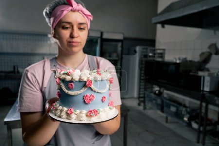 Foto de En una cocina profesional una pastelera está de pie con un pastel terminado en sus manos - Imagen libre de derechos