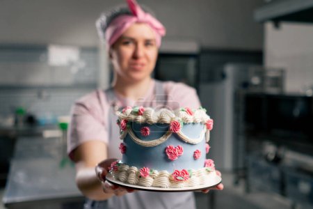 Téléchargez les photos : Dans une cuisine professionnelle une boulangère se tient debout avec un gâteau fini dans ses mains elle sourit - en image libre de droit
