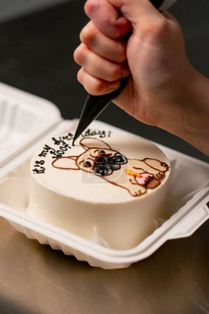 Téléchargez les photos : Gros plan d'une boulangère dans une cuisine professionnelle dessinant un chien sur un gâteau à la crème - en image libre de droit