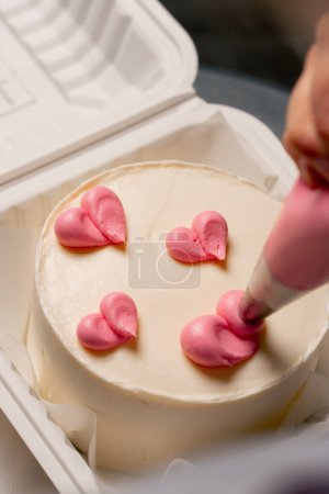 Téléchargez les photos : Gros plan d'une boulangère dans une cuisine professionnelle dessinant un cœur sur un gâteau à la crème rose - en image libre de droit