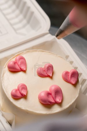 Téléchargez les photos : Gros plan d'une boulangère dans une cuisine professionnelle dessinant un cœur sur un gâteau à la crème rose - en image libre de droit