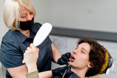 Téléchargez les photos : Cabinet dentaire médecin dentiste blonde consultant un patient regardant autour de la bouche le patient regarde dans le miroir - en image libre de droit