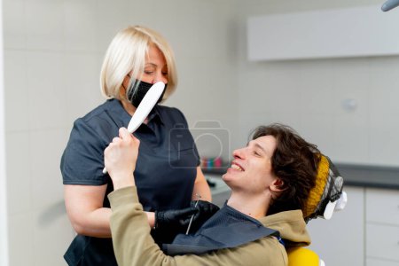Foto de Odontólogo médico odontólogo rubio consulta a un paciente mirando alrededor de la boca el paciente se mira en el espejo - Imagen libre de derechos